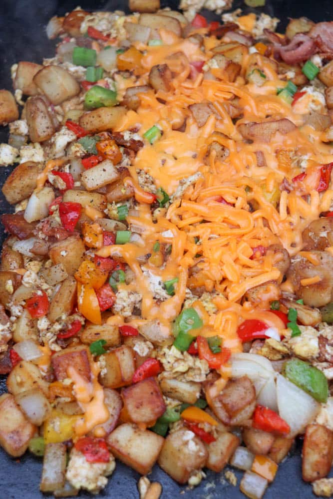 Diced potatoes, bell peppers, onions, eggs, and bacon cooked together on a griddle, topped with shredded melted cheddar cheese and chopped green onions.