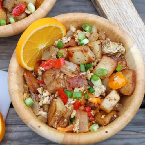 A wooden bowl filled with a breakfast hash of diced potatoes, scrambled eggs, red and yellow bell peppers, onions, and green onions, garnished with an orange slice, on a rustic wooden table.