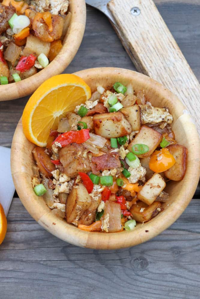 A wooden bowl filled with a breakfast hash of diced potatoes, scrambled eggs, red and yellow bell peppers, onions, and green onions, garnished with an orange slice, on a rustic wooden table.