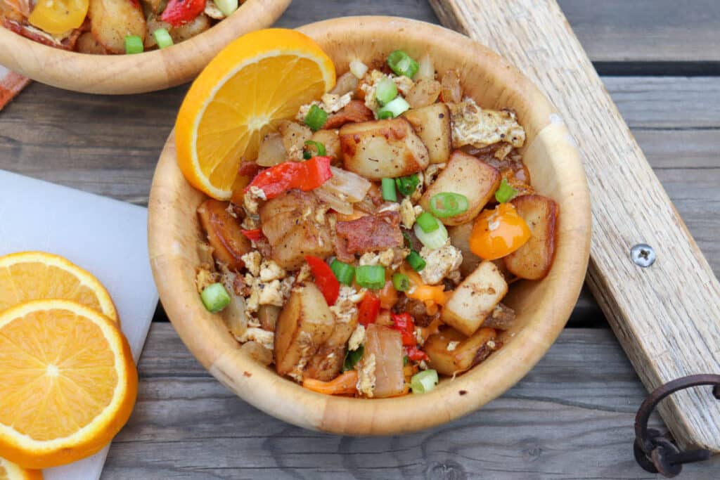 A wooden bowl filled with a breakfast hash of diced potatoes, red bell peppers, onions, scrambled eggs, and green onions, garnished with an orange wedge. Slices of orange are on the side, atop a wooden table.