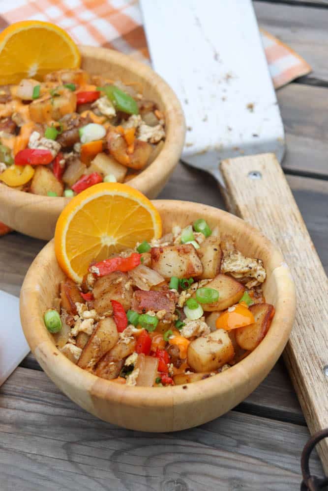Two wooden bowls filled with breakfast hash made of potatoes, peppers, onions, scrambled eggs, and green onions, each garnished with an orange slice. A spatula and checked napkin are nearby on a wooden table.