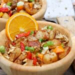 A wooden bowl filled with a colorful potato hash, including diced potatoes, bell peppers, scrambled eggs, and green onions, garnished with a slice of orange. Another similar bowl is in the background.
