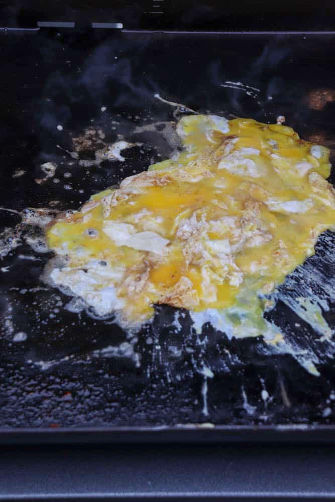 Scrambled eggs cooking on a hot, greased griddle with steam rising, yolk and whites partially mixed and spreading across the dark surface.