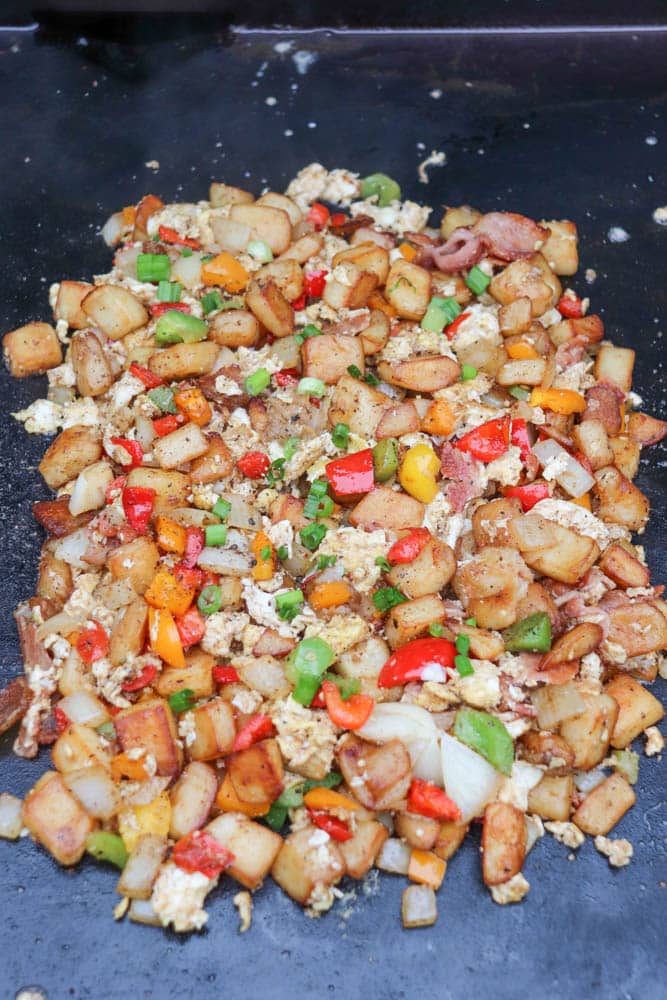 A colorful breakfast hash of diced potatoes, bell peppers, onions, and scrambled eggs cooking on a flat top griddle.