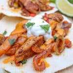 Two flour tortillas topped with seasoned grilled chicken strips, sautéed onions and peppers, melted cheese, sour cream, and cilantro, served with lime wedges on a wooden surface.
