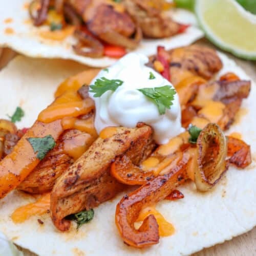 Two flour tortillas topped with seasoned grilled chicken strips, sautéed onions and peppers, melted cheese, sour cream, and cilantro, served with lime wedges on a wooden surface.