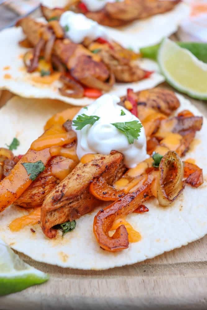 Two flour tortillas topped with seasoned grilled chicken strips, sautéed onions and peppers, melted cheese, sour cream, and cilantro, served with lime wedges on a wooden surface.