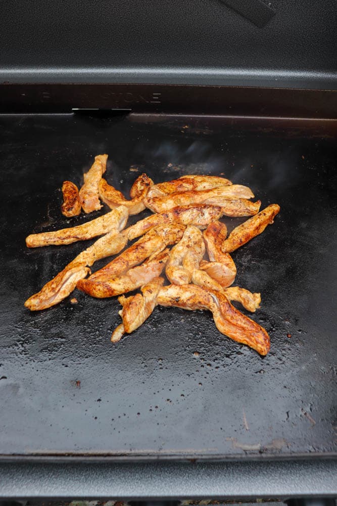 Seasoned strips of chicken cooking on a flat-top grill, with a dark, oily surface and steam rising slightly from the meat.