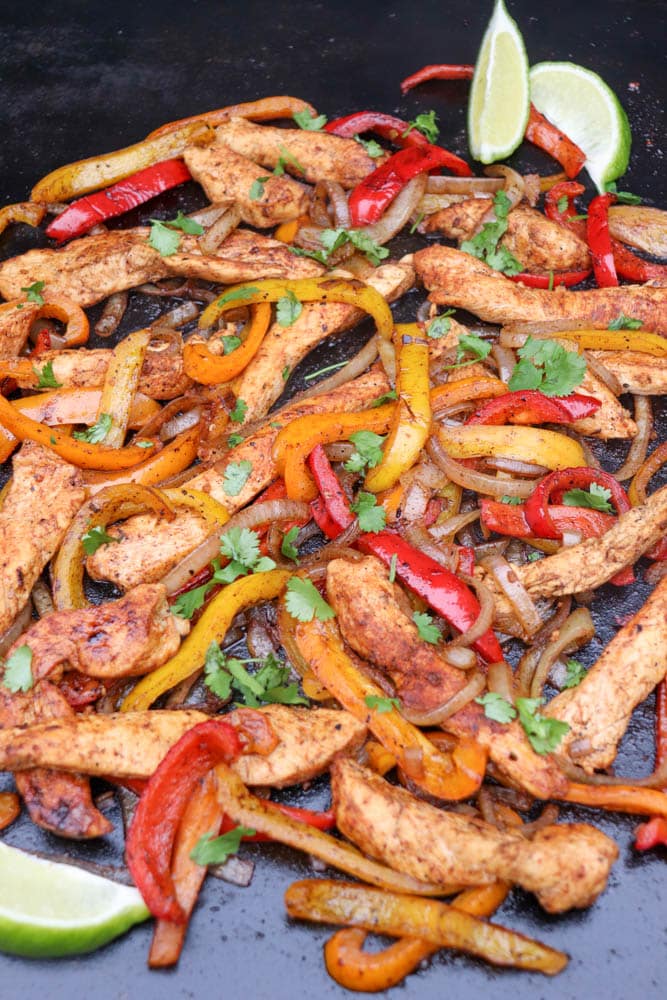 Colorful chicken fajitas with strips of seasoned chicken, red and yellow bell peppers, onions, and fresh cilantro, served on a dark surface with lime wedges in the corner.