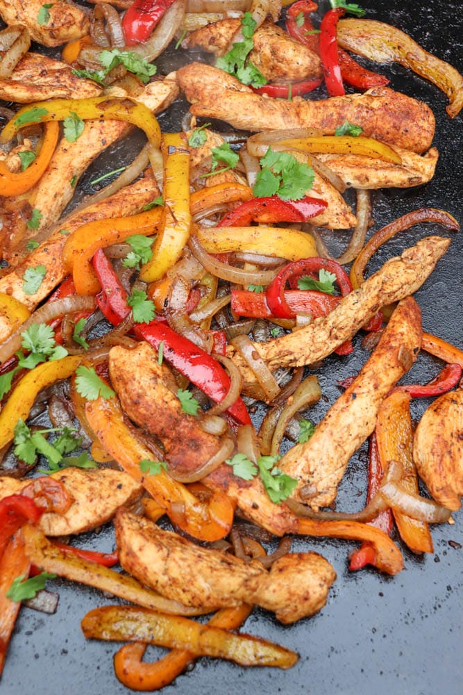 Colorful chicken fajitas with sliced seasoned chicken, red and yellow bell peppers, onions, and fresh cilantro, cooked on a flat griddle. The ingredients are glistening and vibrant.