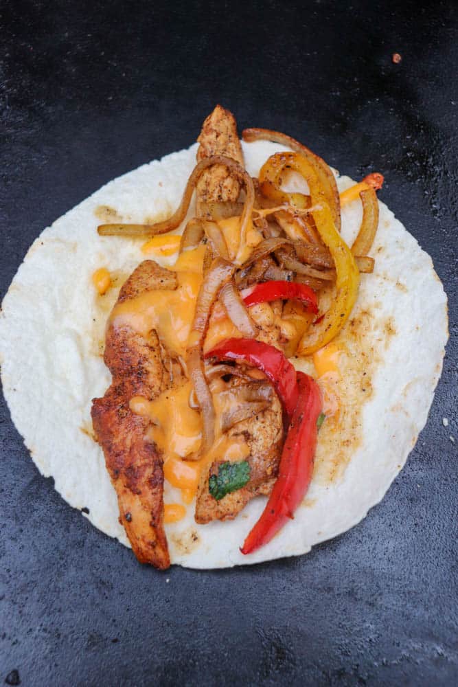 A flour tortilla topped with grilled chicken strips, sautéed onions, red and yellow bell peppers, and melted cheese on a dark surface.