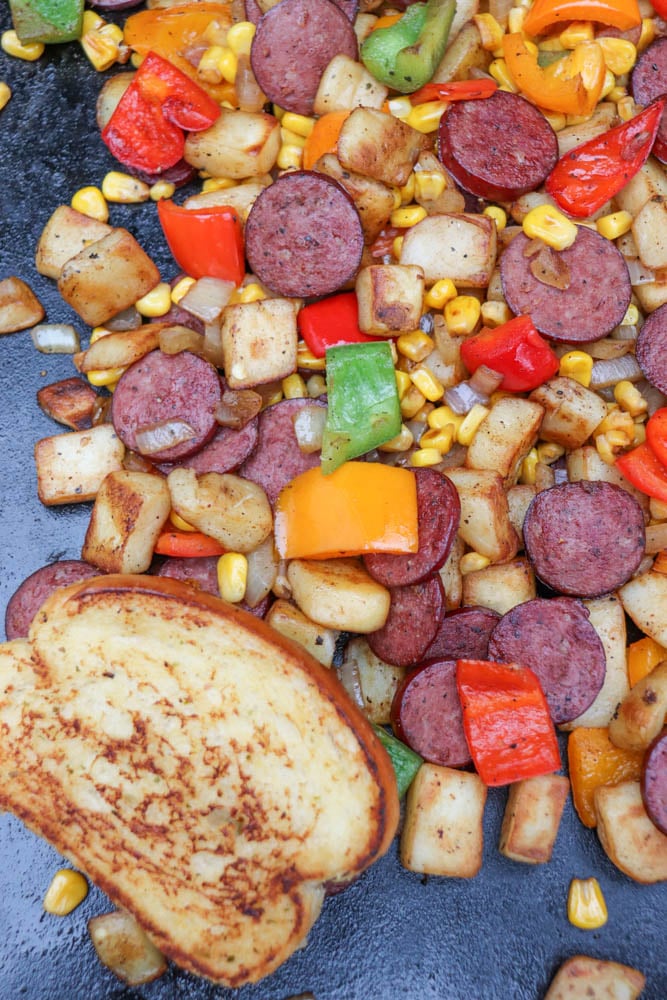 Sliced sausage, diced potatoes, bell peppers, corn, and onions are cooked together on a skillet. A slice of toasted bread is also visible in the bottom left corner.