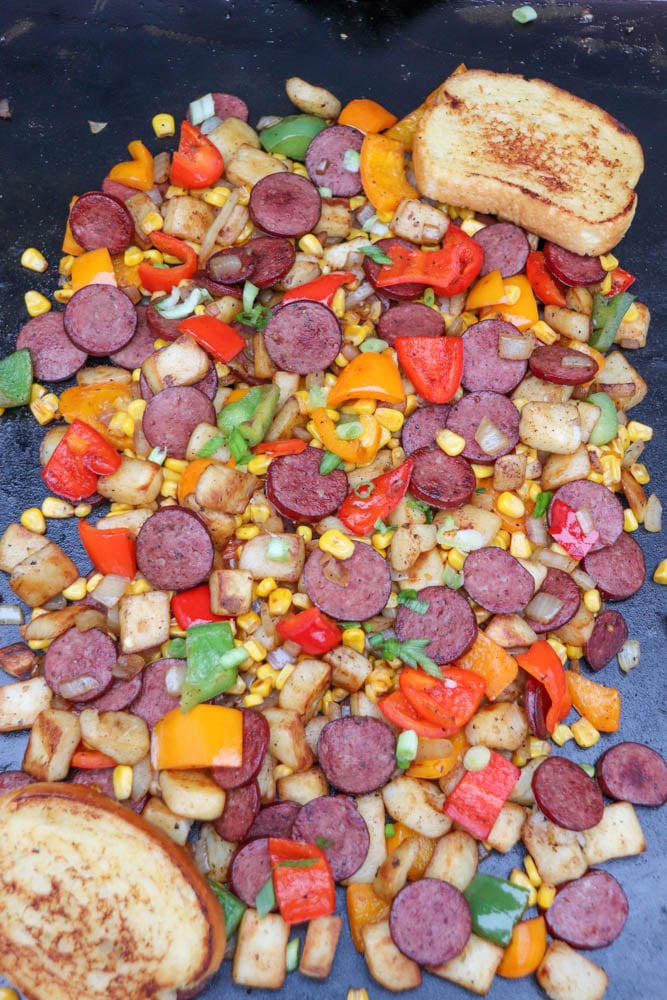A colorful mix of cooked sausage slices, potatoes, bell peppers, corn, and chopped green onions on a grill or griddle, with two slices of toasted bread on the side.