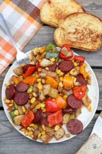 A plate of sautéed sausage slices, diced potatoes, bell peppers, onions, and corn sits on a wooden table beside two slices of toasted bread and a checkered cloth napkin. A fork rests on the plate.