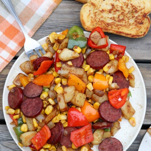 A plate of sautéed sausage slices, diced potatoes, bell peppers, onions, and corn sits on a wooden table beside two slices of toasted bread and a checkered cloth napkin. A fork rests on the plate.