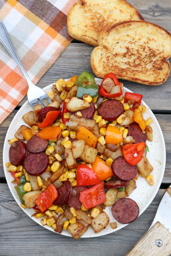 A plate of sautéed sausage slices, diced potatoes, bell peppers, onions, and corn sits on a wooden table beside two slices of toasted bread and a checkered cloth napkin. A fork rests on the plate.