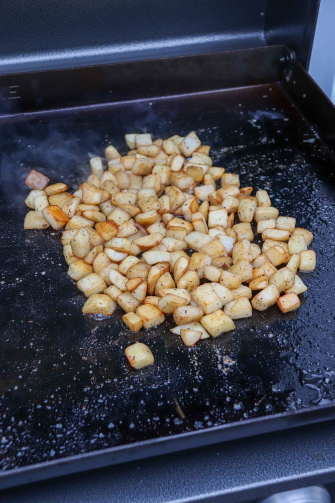 Diced potatoes are being cooked and browned on a flat-top griddle, seasoned with pepper and other spices. Steam rises from the hot surface.