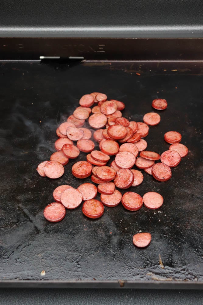 Sliced sausage being cooked on a flat, black griddle, with some pieces lightly browned and a bit of steam rising.