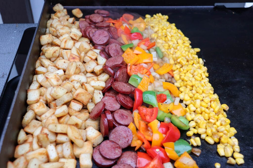 A grill with rows of diced potatoes, sliced sausage, chopped bell peppers in various colors, and yellow corn, all cooking side by side.