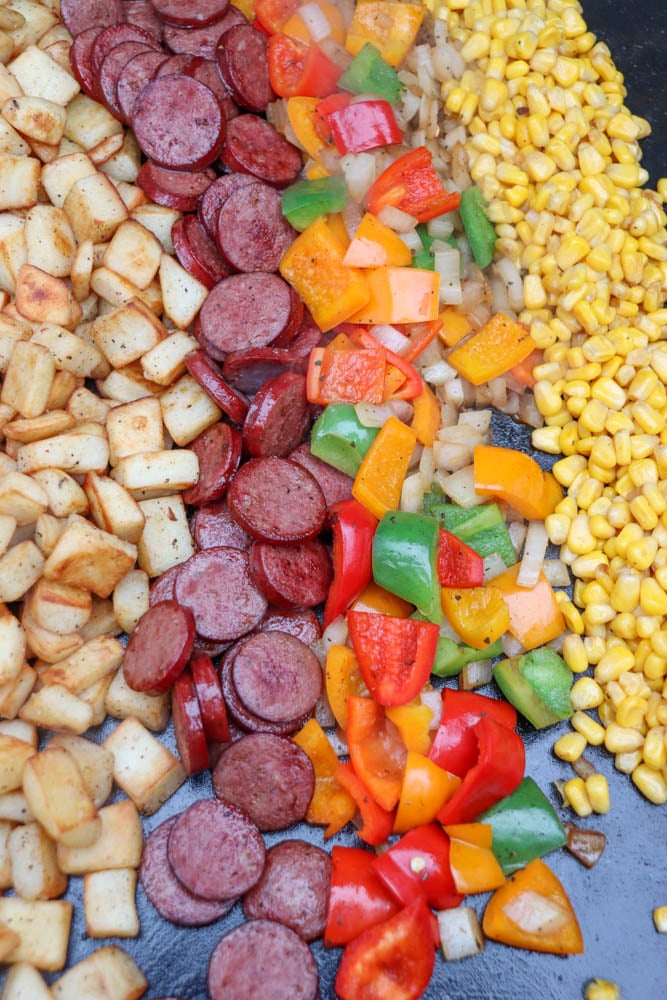 Rows of cooked diced potatoes, sliced sausage, chopped colorful bell peppers, onions, and yellow corn arranged neatly side by side on a flat cooking surface.