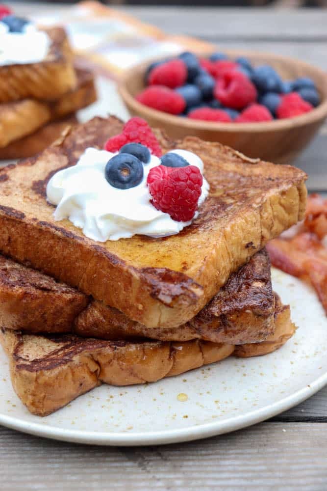 A plate with two slices of French toast topped with whipped cream, blueberries, and raspberries. In the background, there is a bowl of mixed fresh berries and some strips of cooked bacon.