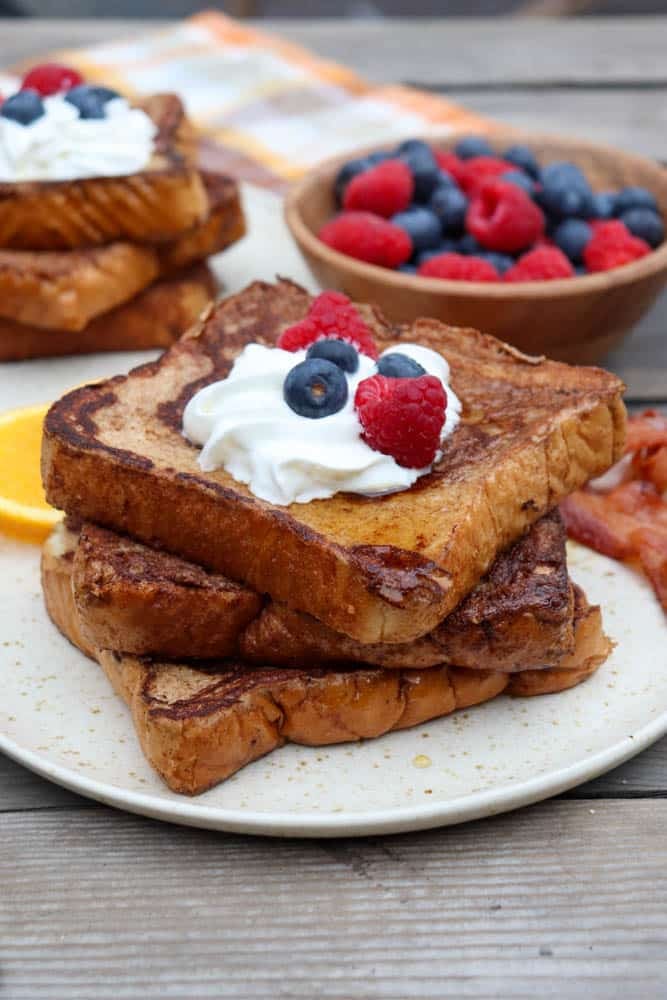 A plate of stacked French toast topped with whipped cream, blueberries, and raspberries, with a slice of orange, bacon, and a bowl of mixed berries in the background.