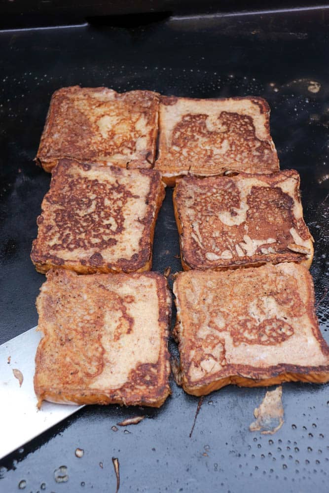 Six slices of golden-brown French toast are cooking on a black griddle. One slice is being lifted slightly with a metal spatula from the lower left corner.