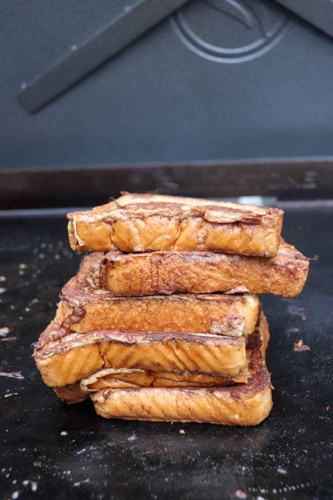 A stack of five golden-brown slices of French toast sits on a dark griddle, with slight charring visible on the edges and a blurred background.