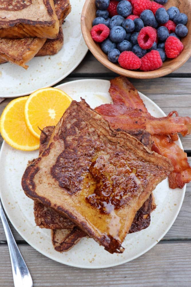 A plate of French toast with syrup, crispy bacon, and orange slices. In the background, a bowl of fresh blueberries and raspberries, and another plate with more French toast are visible on a wooden table.