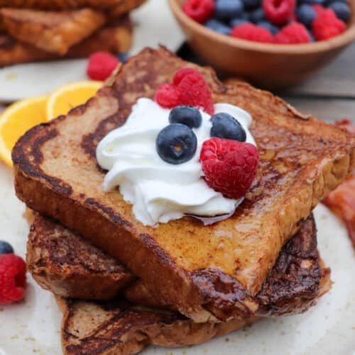 Two thick slices of French toast topped with whipped cream, blueberries, and raspberries on a plate, with more French toast and a bowl of mixed berries in the background.