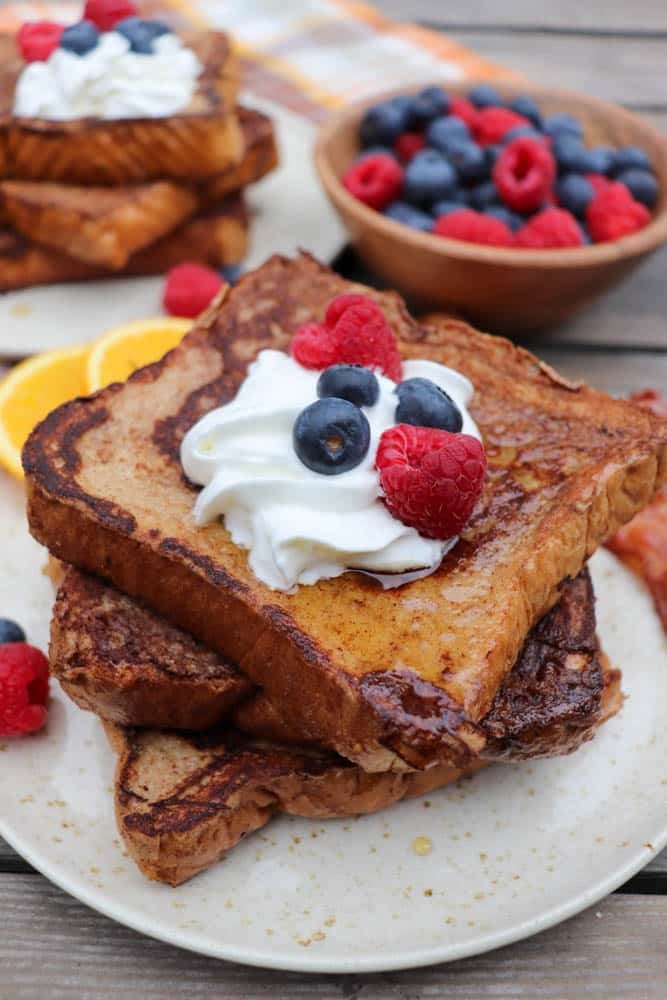 Two thick slices of French toast topped with whipped cream, blueberries, and raspberries on a plate, with more French toast and a bowl of mixed berries in the background.