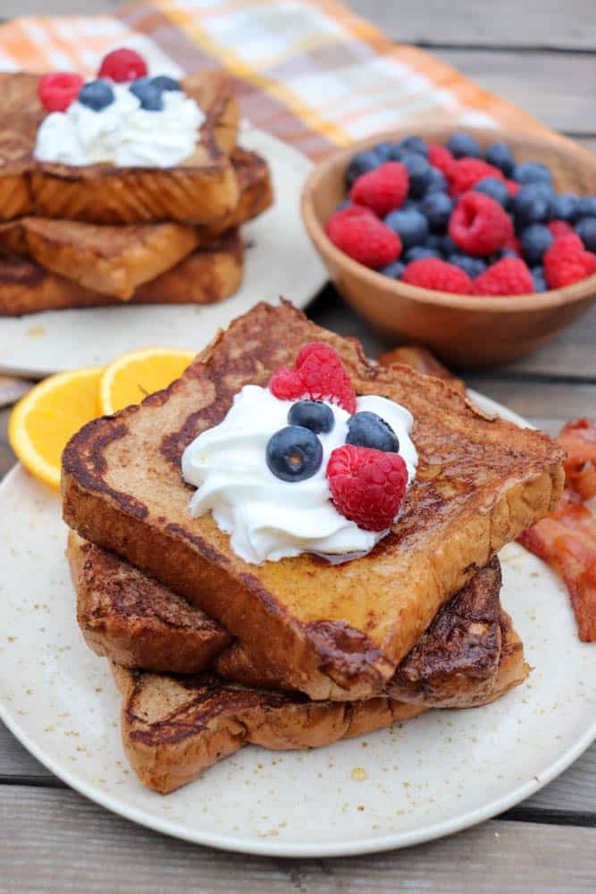 Two slices of French toast topped with whipped cream, raspberries, and blueberries on a plate, with orange slices and bacon on the side. In the background are more French toast and a bowl of mixed berries.