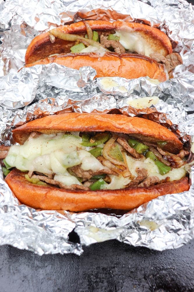 Two Philly cheesesteak sandwiches filled with beef, melted cheese, and bell peppers, served in foil wrappers on a dark surface.