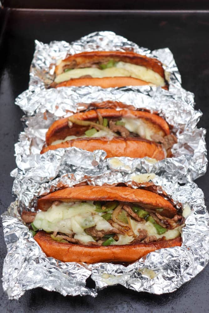 Three cheesesteak sandwiches with melted cheese, grilled onions, and peppers are wrapped in foil and arranged in a row on a dark surface.