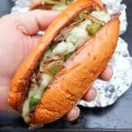A hand holds a toasted hoagie roll filled with melted cheese, sautéed onions, green peppers, and sliced beef, resembling a Philly cheesesteak. Another sandwich wrapped in foil is visible in the background.