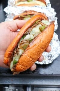 A hand holds a toasted hoagie roll filled with melted cheese, sautéed onions, green peppers, and sliced beef, resembling a Philly cheesesteak. Another sandwich wrapped in foil is visible in the background.