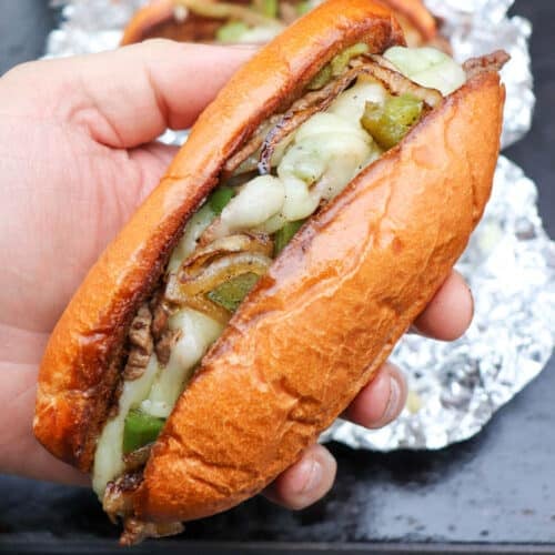 A hand holds a toasted hoagie roll filled with melted cheese, sautéed onions, green peppers, and sliced beef, resembling a Philly cheesesteak. Another sandwich wrapped in foil is visible in the background.
