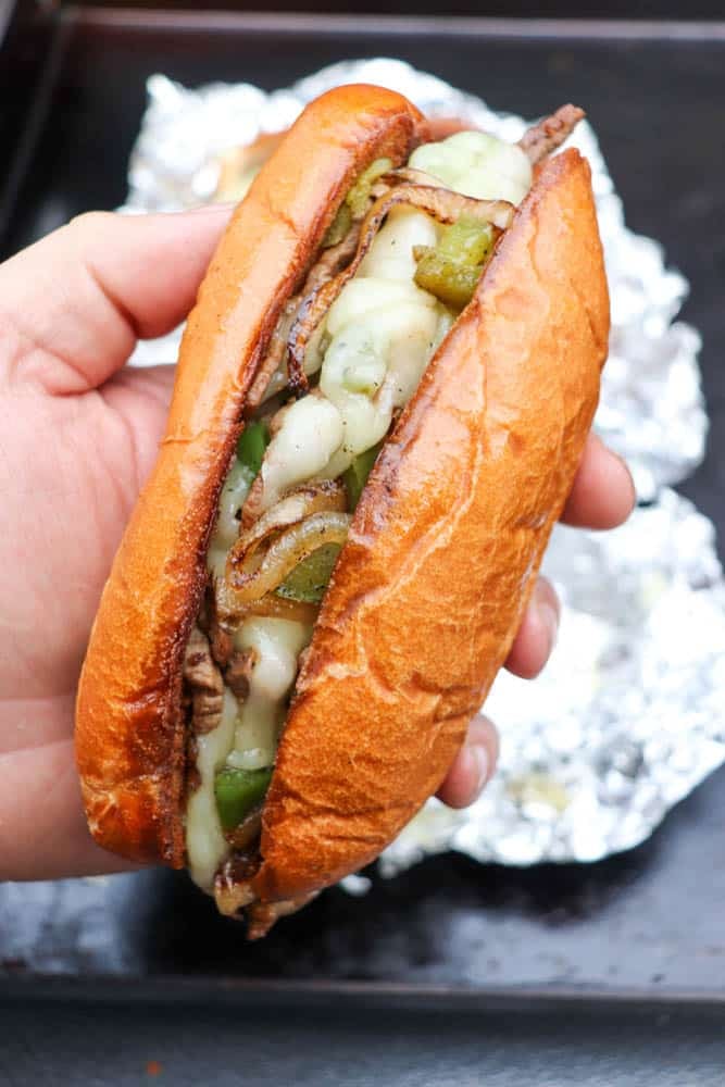 A hand holding a hoagie roll filled with sliced beef, melted cheese, sautéed onions, and green bell peppers. Foil is visible in the background on a dark surface.