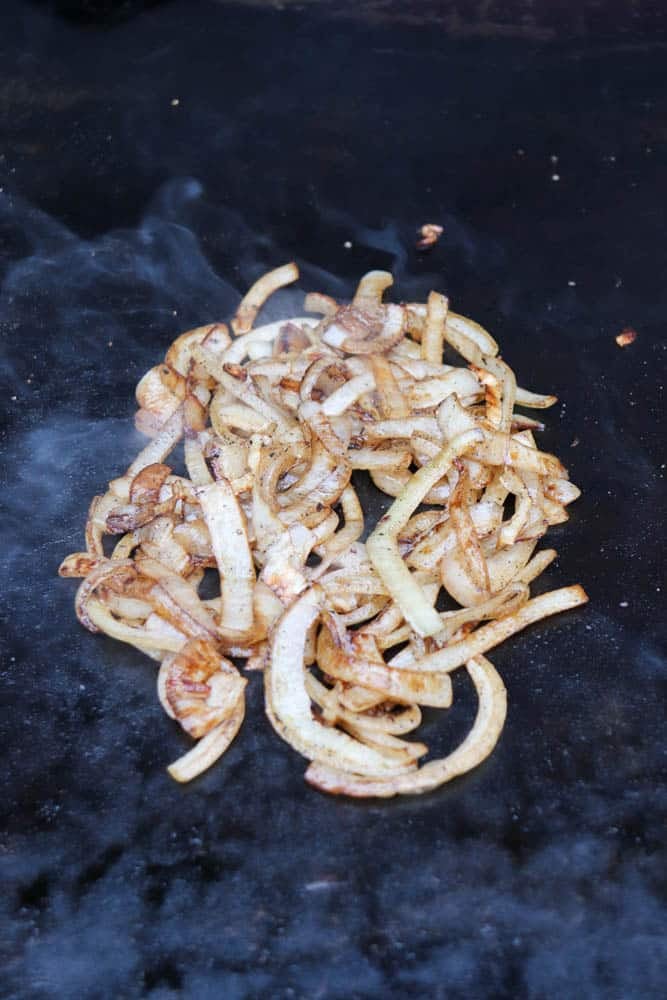 Sliced onions being cooked and caramelized on a hot, flat grill with visible steam rising around them.