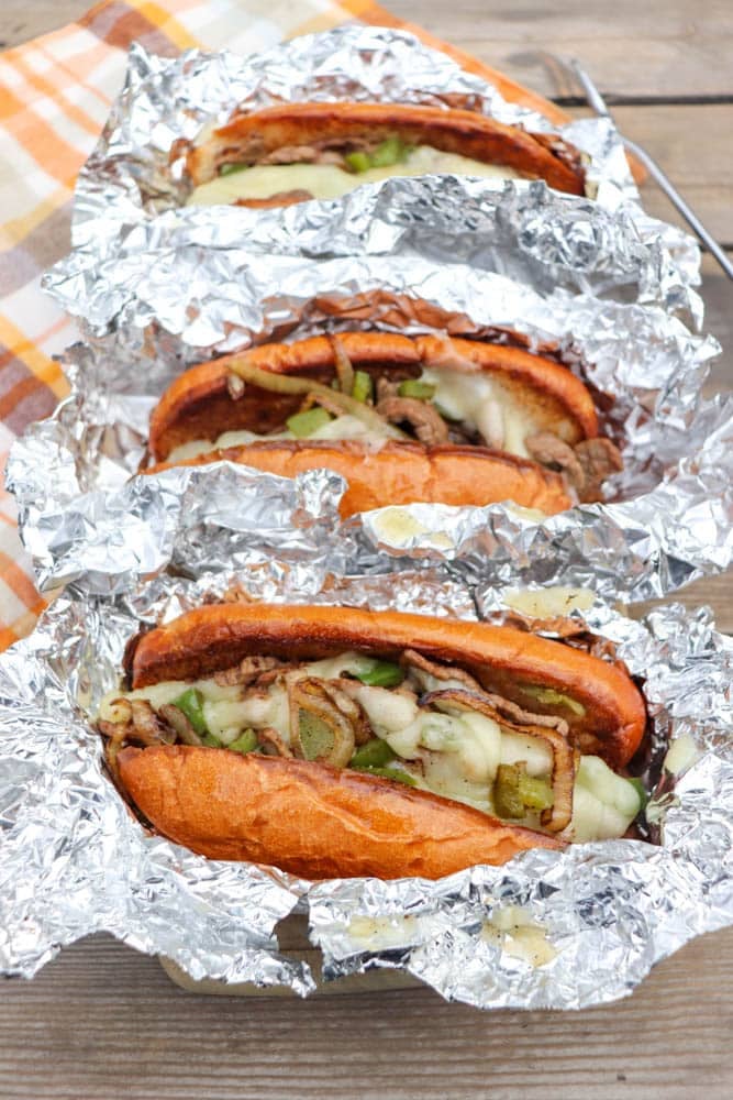Three cheesesteak sandwiches filled with steak, melted cheese, and green peppers are wrapped in foil and set on a wooden surface with a plaid napkin nearby.