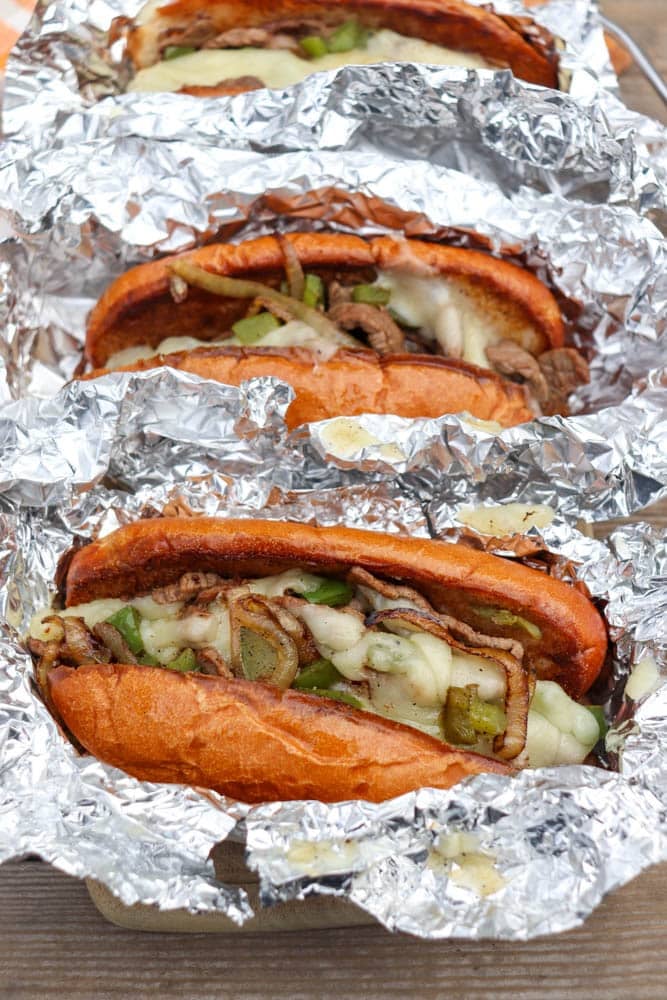 Three cheesesteak sandwiches in foil, filled with melted cheese, sautéed onions, and green peppers on toasted hoagie rolls.