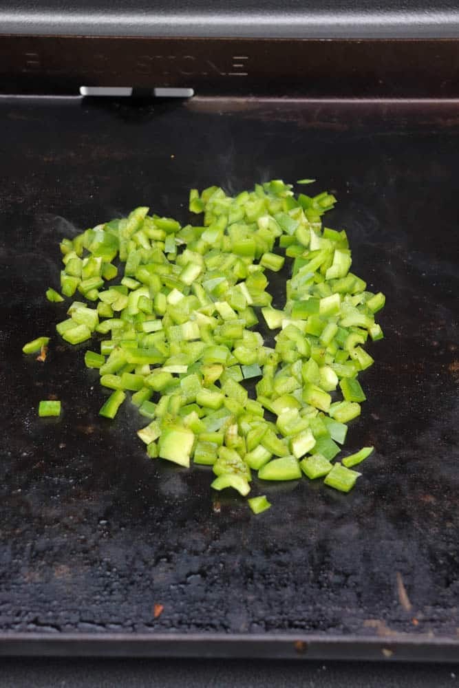 Diced green bell peppers cooking on a flat, black griddle surface, with slight steam rising from the vegetables.