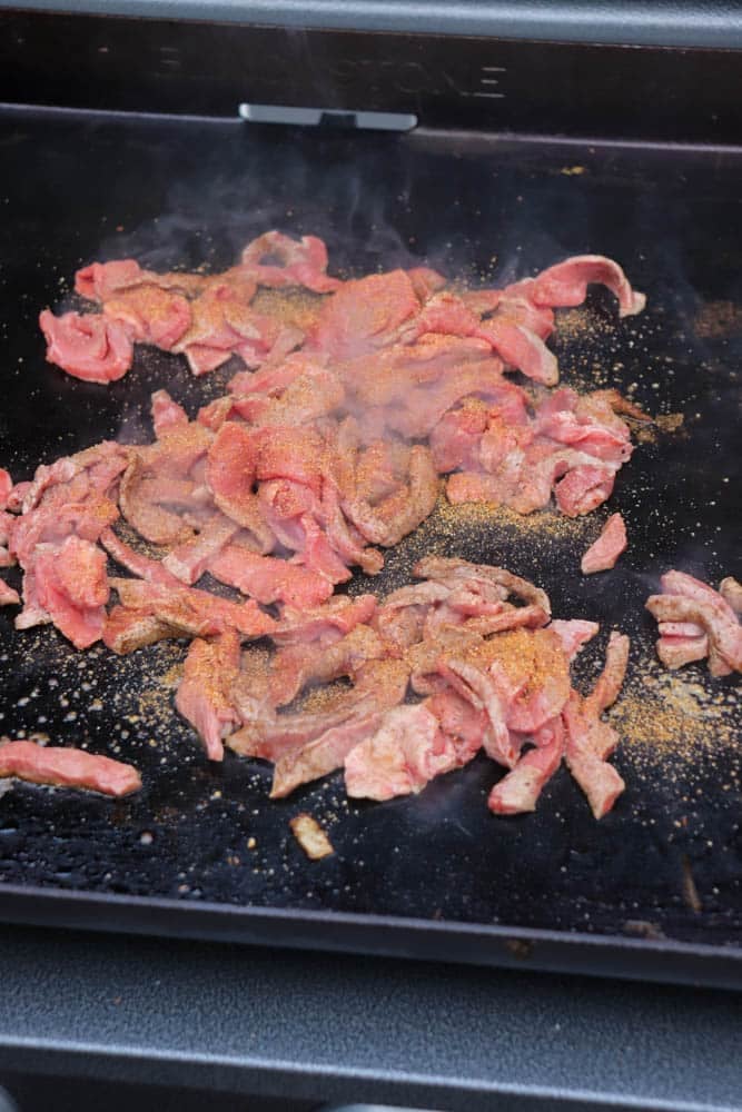 Thin slices of raw beef, seasoned with spices, are cooking on a hot flat-top grill, with steam rising from the meat.
