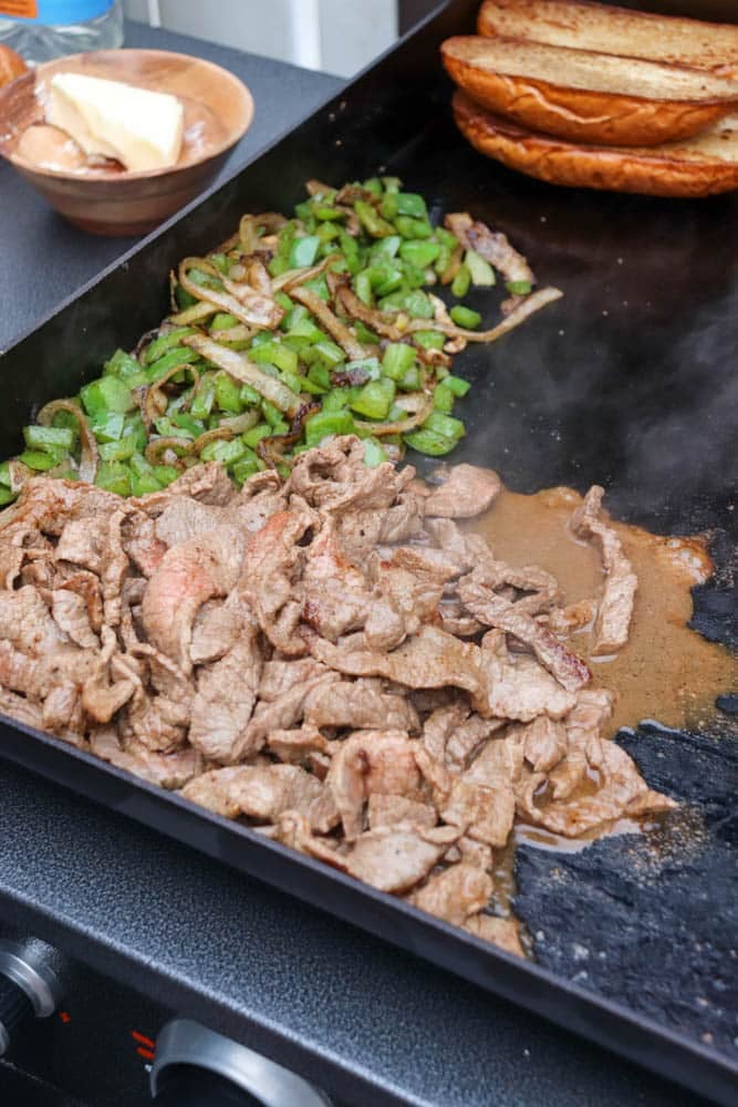 Thin slices of cooked steak, sautéed green bell peppers and onions, and toasted sandwich rolls are cooking on a flat-top griddle, with a dish of butter nearby.