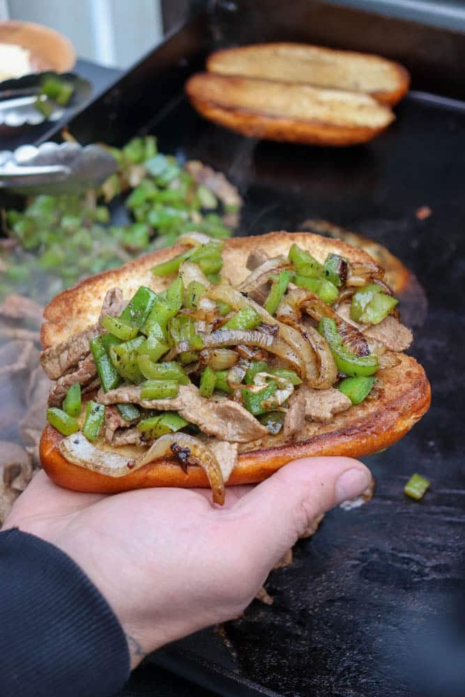 A hand holds a sandwich filled with sliced beef, sautéed green peppers, and onions, with more ingredients and a toasted bun on a grill in the background.