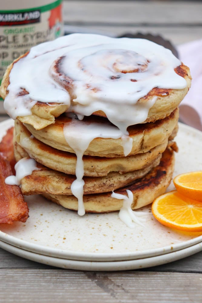 A stack of cinnamon roll pancakes topped with white icing is served on a plate with crispy bacon and orange slices. A jar of organic maple syrup is partially visible in the background.