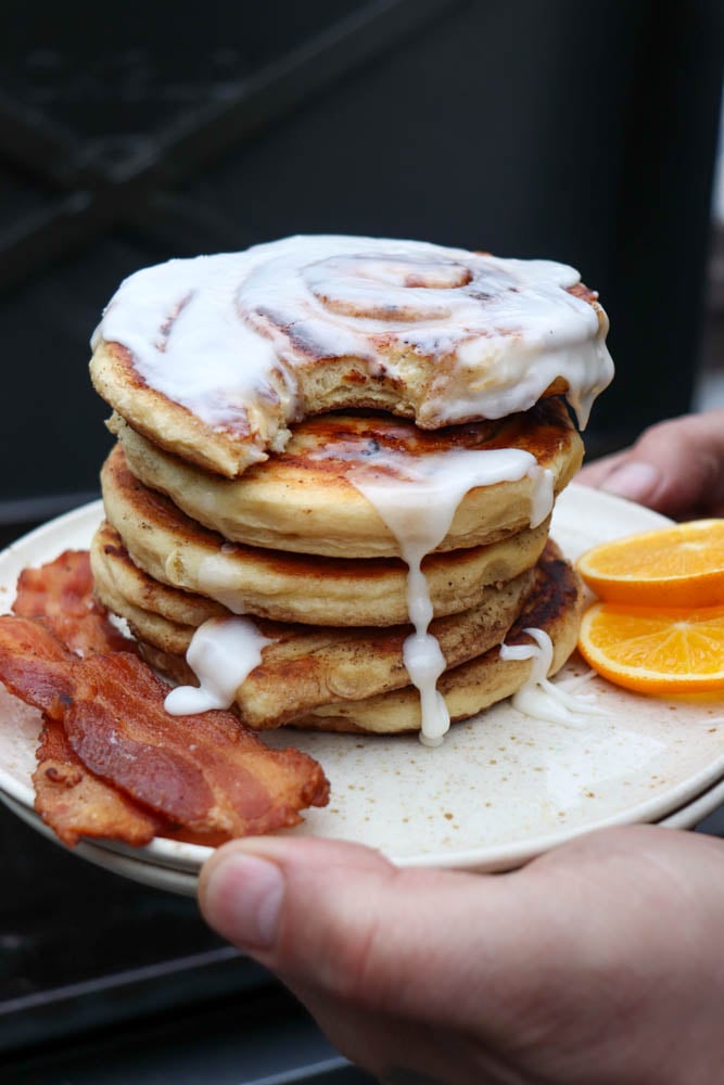 A hand holds a plate with a stack of pancakes topped with white icing, two strips of crispy bacon, and orange slices on the side.