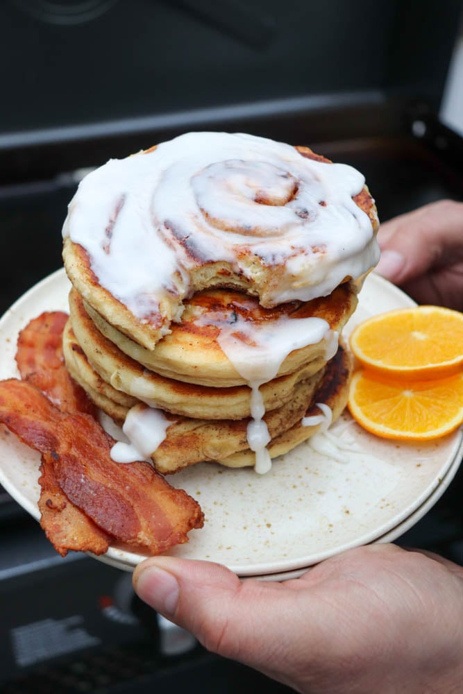 A plate with a stack of cinnamon roll pancakes topped with white icing, two strips of crispy bacon, and two orange slices, being held in someone’s hands.