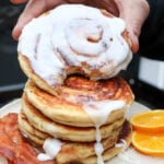 A hand holds a cinnamon roll pancake with icing, stacked on top of more pancakes. On the plate are crispy bacon strips and orange slices. Icing drips down the pancakes.