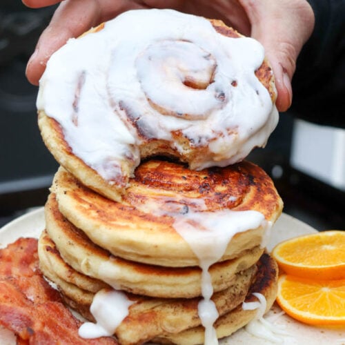 A hand holds a cinnamon roll pancake with icing, stacked on top of more pancakes. On the plate are crispy bacon strips and orange slices. Icing drips down the pancakes.